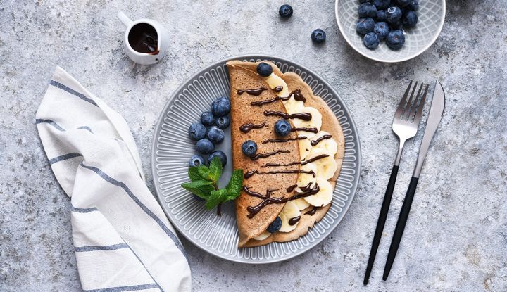 Schwester Bernadettes Pfannkuchen mit Gerste schmecken lecker nussig mit Bananenscheiben, frischen Blaubeeren und Schokosoße, angerichtet auf einem grauen Teller mit Minze, daneben Besteck und Serviette, Pfannkuchen mit Gerste