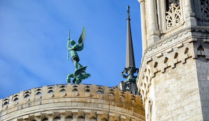 Erzengel Michael: Mit der Lanze gegen das Böse Statue des Erzengels Michael mit Schwert und Fahne auf dem Dach einer Kirche, neben einem Kirchturm mit filigranen Verzierungen vor blauem Himmel
